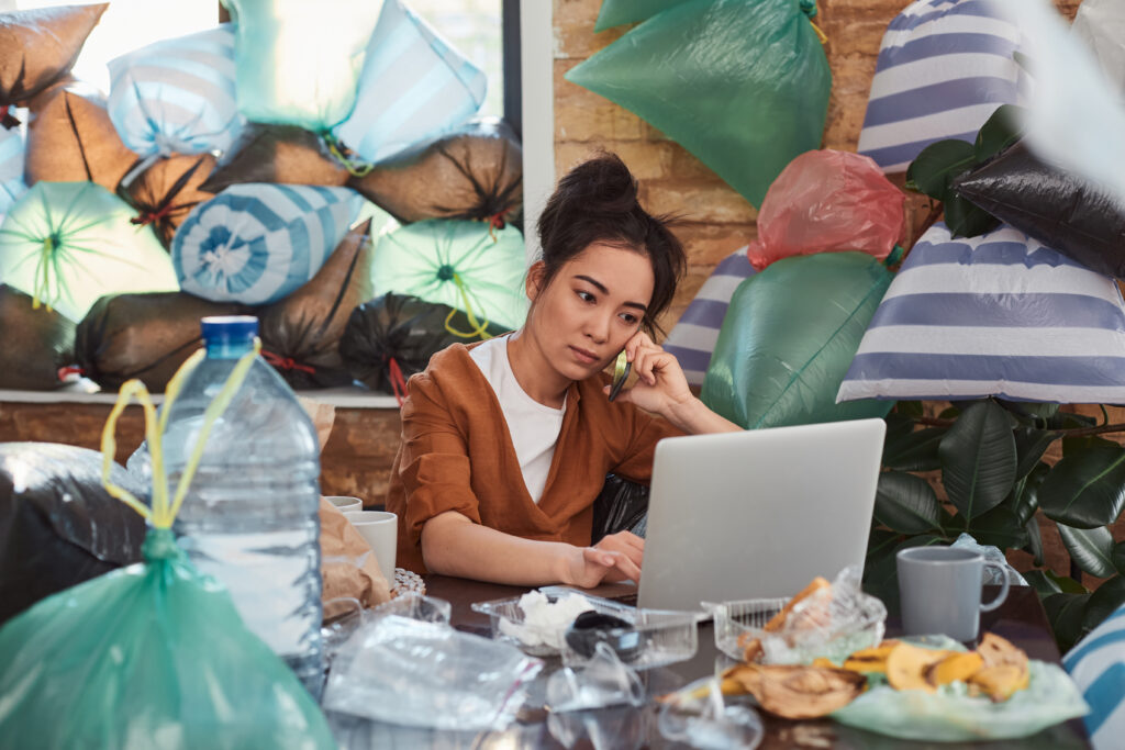 woman on the phone working on her laptop as she sits in her hoarder home with trash all around her